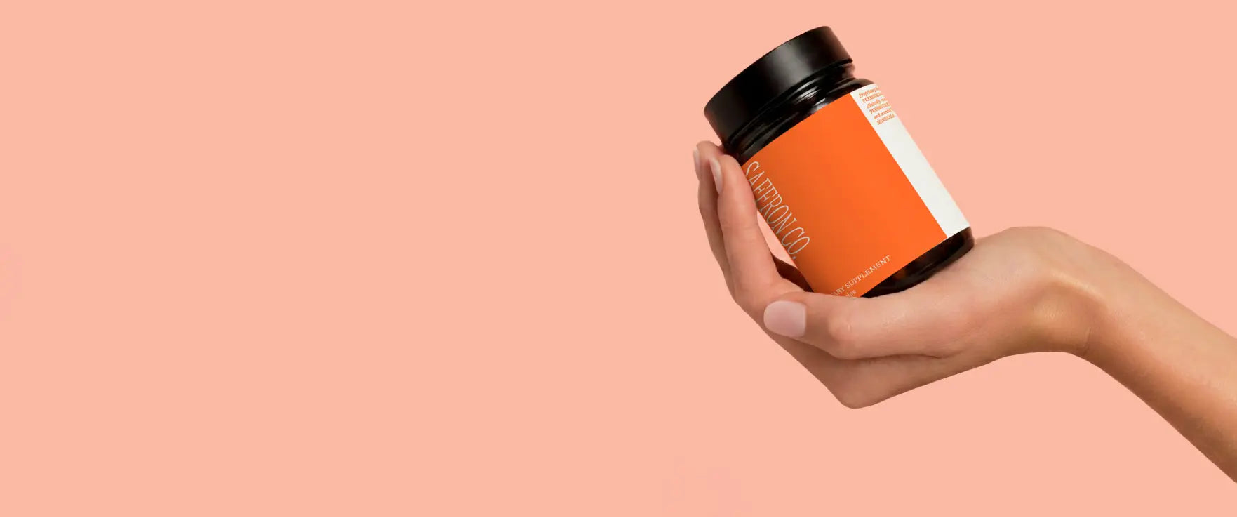 Hand holding a supplement jar with an orange label against a pink background